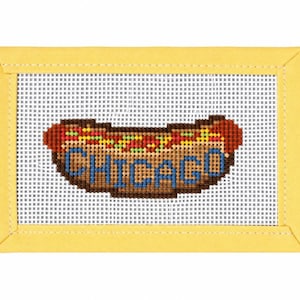 May include: A rectangular needlepoint canvas featuring a hot dog design with the word CHICAGO stitched in blue thread. The hot dog is brown with red and green accents, centered on a white grid mesh, and framed by a solid yellow fabric border.