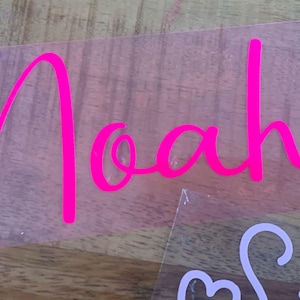 Op de afbeelding: Een roze vinyl sticker met de naam "Noah" geschreven in een cursief schrift.