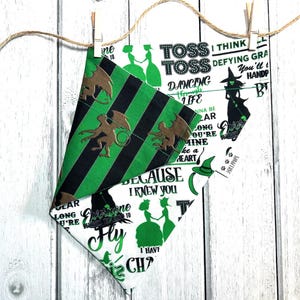 Può includere: Un tessuto a strisce verde e nero con un design di strega e creatura. Il tessuto è piegato a forma di triangolo e ha il testo che recita "Toss Toss Dancing Through Life I Think Defying Gravity You'll Hand Pick Because I Knew You Everyone Fly I Have A Witchy Charm".