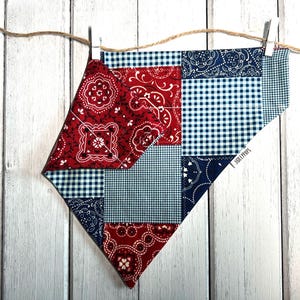 May include: A red, white and blue bandana with a patchwork design. The bandana is made of cotton fabric and has a frayed edge. It is hanging on a white wooden background.