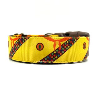 May include: A yellow dog collar with a colourful pattern. The collar features a repeating design of orange shapes, a diagonal stripe of colourful dots, and a green tree icon. The collar has a brown plastic buckle.