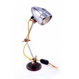 Könnte beinhalten: Eine einzigartige Schreibtischlampe aus recycelten Metallkomponenten. Die Lampe hat einen verchromten Scheinwerfer, einen Schraubenschlüsselarm und einen kreisförmigen Sockel. Das Design umfasst ein gelbes Elektrokabel und einen schwarzen Ein-/Ausschalter, für einen industriellen Look. Höhe: ca. 25 cm.