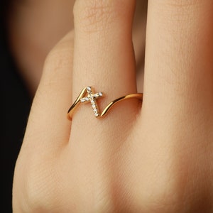 以下が含まれることがあります： ダイヤモンドで飾られた小さな十字架のデザインが施された金の指輪。指輪は指にはめられています。