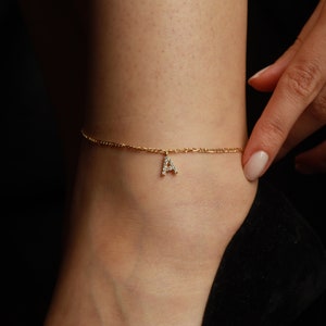 May include: A gold anklet featuring a delicate chain and a sparkling letter "A" pendant. The anklet is worn on an ankle, with a hand gently touching the chain. The background is dark, highlighting the jewellery.