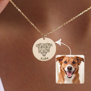 Puede incluir: Un collar dorado con un colgante circular que presenta un retrato grabado detallado de la cara de un perro y el nombre "Kobe". Una pequeña foto del perro se muestra junto al colgante.