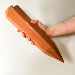 Pode incluir: Um tampão de garrafa em forma de lápis cor de terracota com uma rolha de cortiça. O tampão tem um design geométrico e uma extremidade pontiaguda. O produto é segurado por uma mão.
