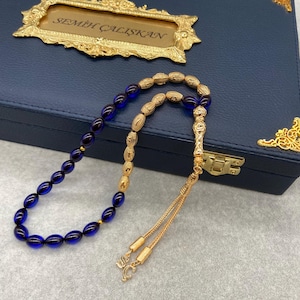 May include: A gold-coloured beaded necklace with dark blue oval beads. The necklace is displayed on a navy blue box with gold accents and a gold-framed plaque that reads "SEMEH ÇALIŞKAN".