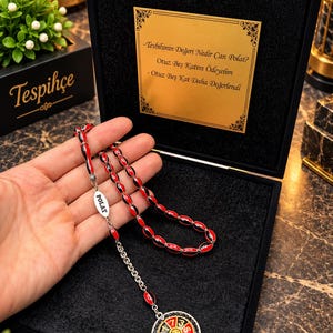 May include: A prayer bead strand featuring red and black oval beads, a silver-toned metal tag engraved with the word POLAT, and a circular pendant with a roulette wheel design. The set is presented in a black box with a gold-colored plaque.