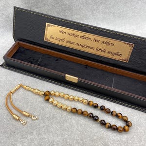 Puede incluir: Un collar de cuentas de oración dorado con cuentas marrones y una borla dorada decorativa, sobre una superficie gris. El collar está junto a una caja negra abierta con una placa dorada con texto.