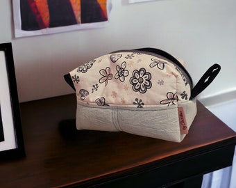 Op de afbeelding: Een beige en grijze toilettas met een bloemenprint, een zwarte rits en een zwart handvat. De tas heeft een label met de tekst "by Crochet"