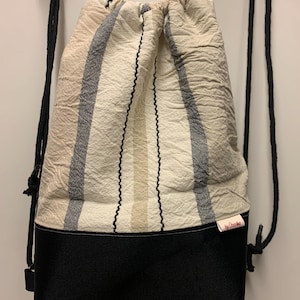 Op de afbeelding: Een wit-zwarte gestreepte rugzak met trekkoord en een zwarte bodem. De tas heeft een klein wit label met de tekst "by Ocoobo" erop.