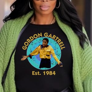 May include: Black t-shirt featuring a circular design with the text "GORDON GARTRELL" in a gold font, surrounding an image of a person in a yellow shirt. The text "Est. 1984" is below the image. The person is wearing a green cardigan.
