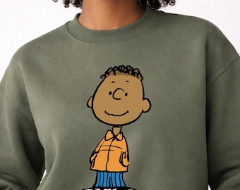 May include: Olive green sweatshirt featuring a cartoon illustration of Franklin from Peanuts. The character is depicted in an orange jacket, blue pants, and white shoes. The name "FRANKLIN" is printed in black at the bottom.