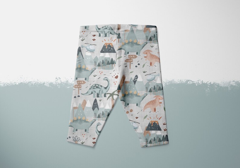 Puede incluir: Leggins grises con estampado de dinosaurios y monta&ntilde;as. El estampado presenta colores azul, verde, marr&oacute;n y naranja. Los leggins tienen un fondo blanco.