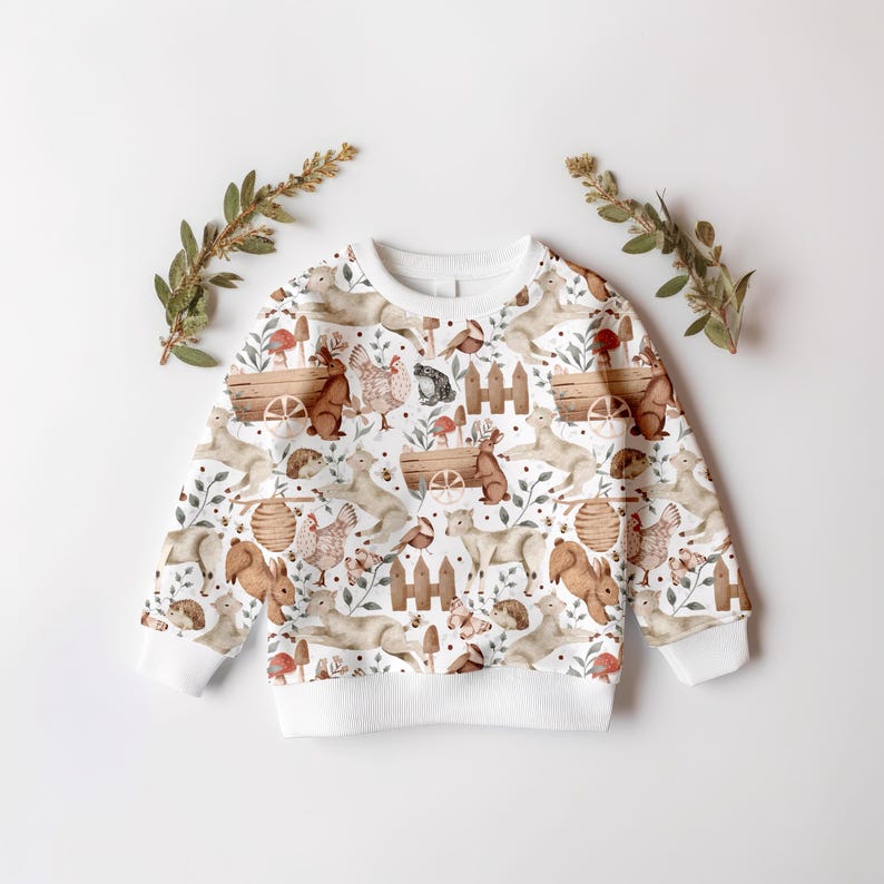 May include: White sweatshirt with a woodland animal print. The print features squirrels, rabbits, birds, and other forest creatures in brown, beige, and green tones.