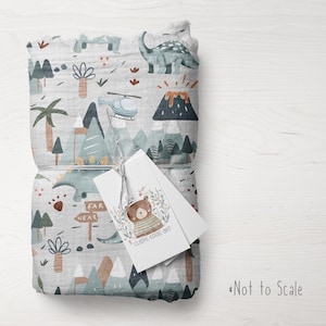 Puede incluir: Una manta de muselina gris y blanca con estampado de dinosaurios, monta&ntilde;as y palmeras. La manta est&aacute; doblada y atada con una etiqueta blanca que dice "Claire Eddie Art" y "Far Near".
