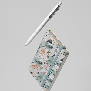 Puede incluir: Una peque&ntilde;a libreta azul p&aacute;lida con un patr&oacute;n de dinosaurios y volcanes. La libreta tiene un cierre el&aacute;stico gris y flota en el aire sobre una pluma blanca.