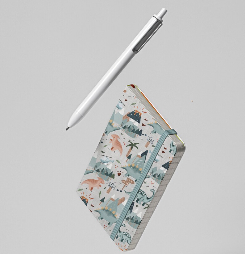 Puede incluir: Una peque&ntilde;a libreta azul p&aacute;lida con un patr&oacute;n de dinosaurios y volcanes. La libreta tiene un cierre el&aacute;stico gris y flota en el aire sobre una pluma blanca.