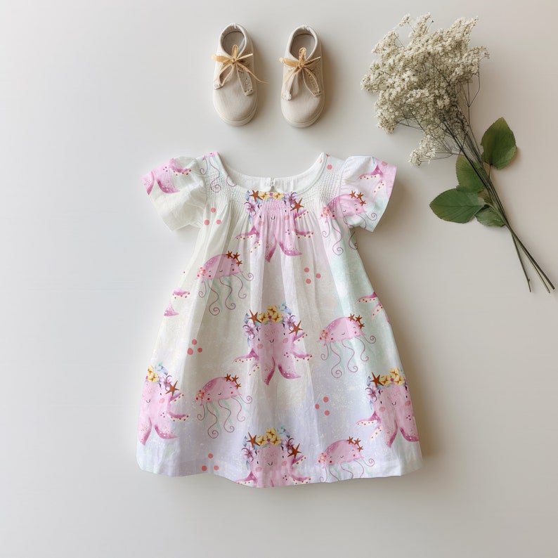 May include: A white dress with short sleeves and a pink octopus print. The dress has a smocked bodice and a gathered skirt. The octopus is wearing a crown of flowers.