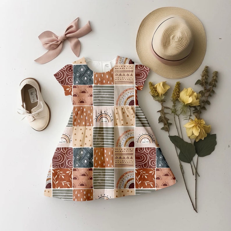 May include: A beige and brown patterned dress with short sleeves and a gathered skirt. The dress features a geometric pattern with sun, rainbow, and abstract shapes.