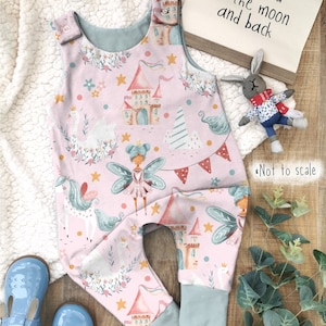May include: Pink baby onesie with a whimsical fairy, unicorn, and castle print. The onesie has a light blue trim and snap closures. The text "love you to the moon and back" is visible in the background.