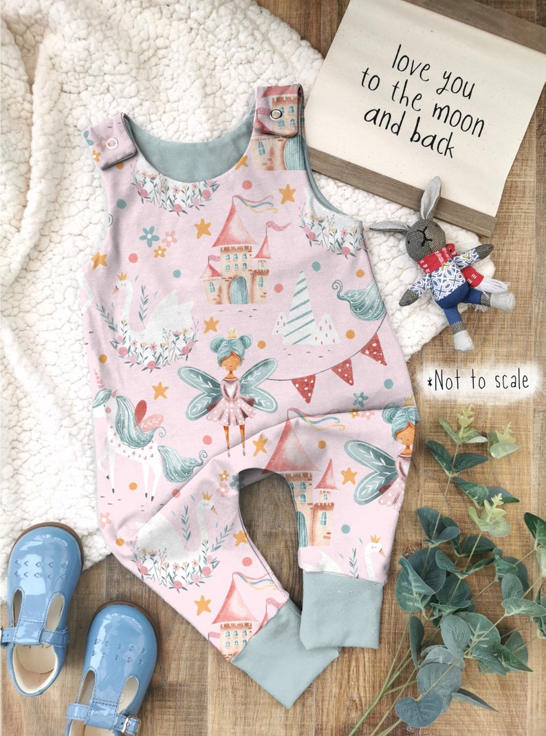 May include: Pink baby onesie with a whimsical fairy, unicorn, and castle print. The onesie has a light blue trim and snap closures. The text "love you to the moon and back" is visible in the background.