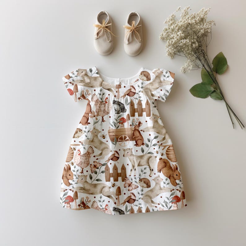 May include: A white dress with a woodland animal print. The dress has short sleeves and a gathered bodice. The print features squirrels, rabbits, birds, and other woodland creatures.