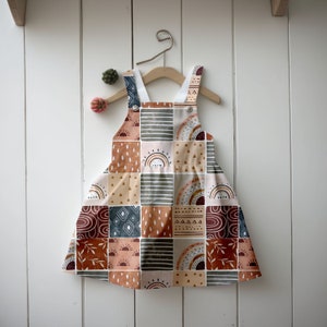 May include: A patchwork-style baby overall dress with a geometric pattern in shades of brown, orange, beige, and blue. The dress features a rainbow, sun, and leaf motifs.