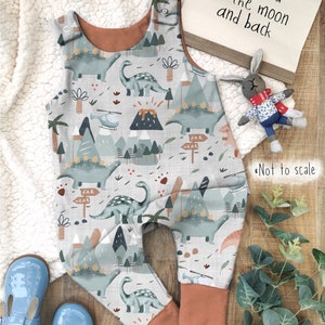Puede incluir: Un pelele de beb&eacute; gris y blanco con estampado de dinosaurios y monta&ntilde;as. El pelele tiene tirantes marrones y una cintura marr&oacute;n. El texto "love you to the moon and back" es visible en el fondo.