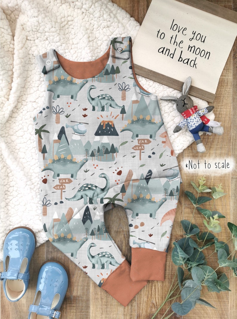 Puede incluir: Un pelele de beb&eacute; gris y blanco con estampado de dinosaurios y monta&ntilde;as. El pelele tiene tirantes marrones y una cintura marr&oacute;n. El texto "love you to the moon and back" es visible en el fondo.