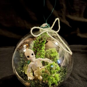May include: A clear glass ornament with a miniature skeleton figure inside, surrounded by green moss and artificial plants. The ornament is suspended by a green wire and a white string bow. The background is dark.