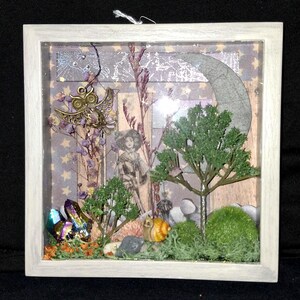 Puede incluir: Una obra de arte enmarcada de técnica mixta que representa una escena de bosque caprichosa. La obra de arte incluye un dije de búho de latón, un recorte de papel de una mujer, árboles en miniatura, musgo, cristales y una luna creciente.