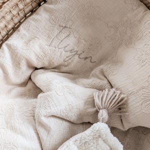 Peut inclure: Une couverture de bébé beige avec des motifs floraux brodés et le nom "Liyin" en écriture cursive. La couverture, ornée d'un pompon, est disposée dans un panier en osier, créant une ambiance douce et chaleureuse.