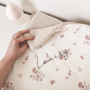 Pode incluir: Um cobertor para bebé branco e bege com um padrão floral e o nome "Louisy" bordado em letras pretas.