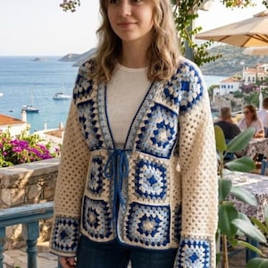 May include: A cream-colored crochet cardigan with blue square patterns. The open-front sweater has long sleeves and a tie closure. The design features a mix of light and dark blue squares, creating a geometric pattern. The cardigan is worn over a white top and blue jeans.