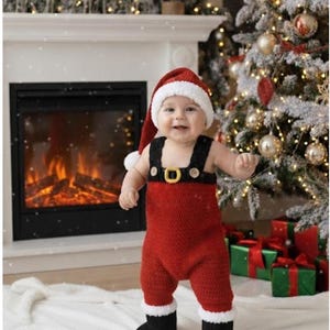 May include: A baby wearing a red Santa Claus outfit, including a hat and jumpsuit with suspenders and a gold buckle. The baby is standing in front of a fireplace and Christmas tree.