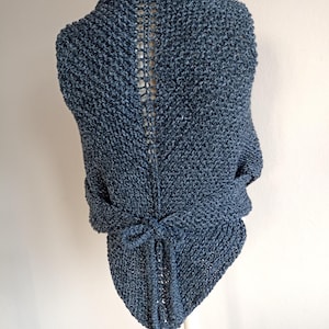 May include: A blue knit wrap with a tie closure. The wrap is displayed on a mannequin, showcasing the back with a detailed open stitch design down the center. The wrap has a textured appearance and a soft, muted color.