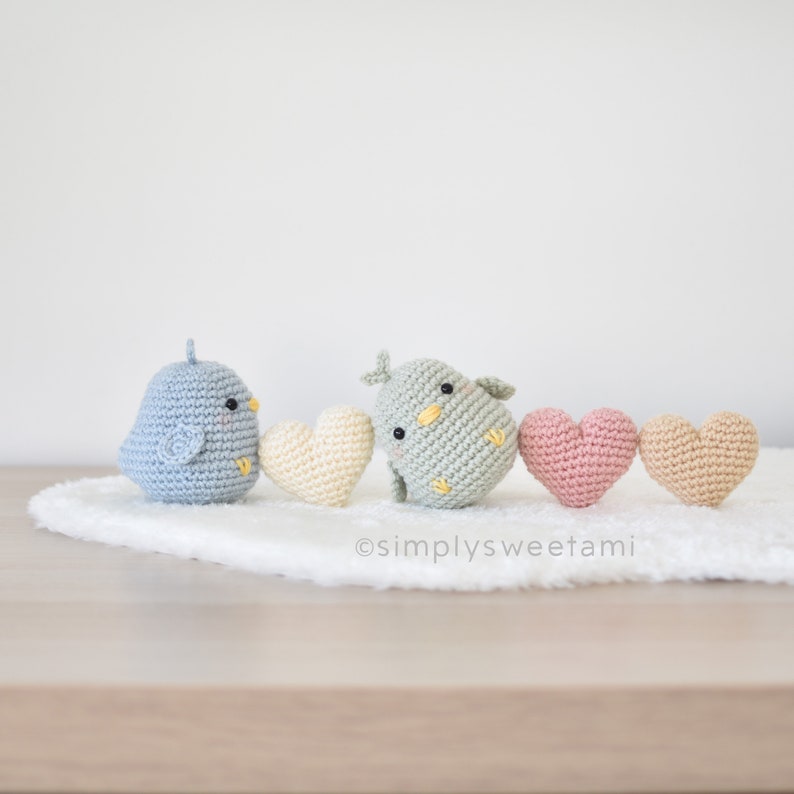 Może przedstawiać: Rząd czterech pluszowych zabawek amigurumi: dwa niebieskie i zielone ptaki oraz dwie zabawki w kształcie serca w kolorze kremowym, r&oacute;żowym i beżowym.