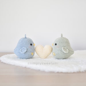 Puede incluir: Dos pájaros de crochet, uno azul claro y otro verde claro, se miran con un corazón de crochet entre ellos. Los pájaros tienen pequeños ojos negros y picos amarillos. El corazón es de color crema claro.
