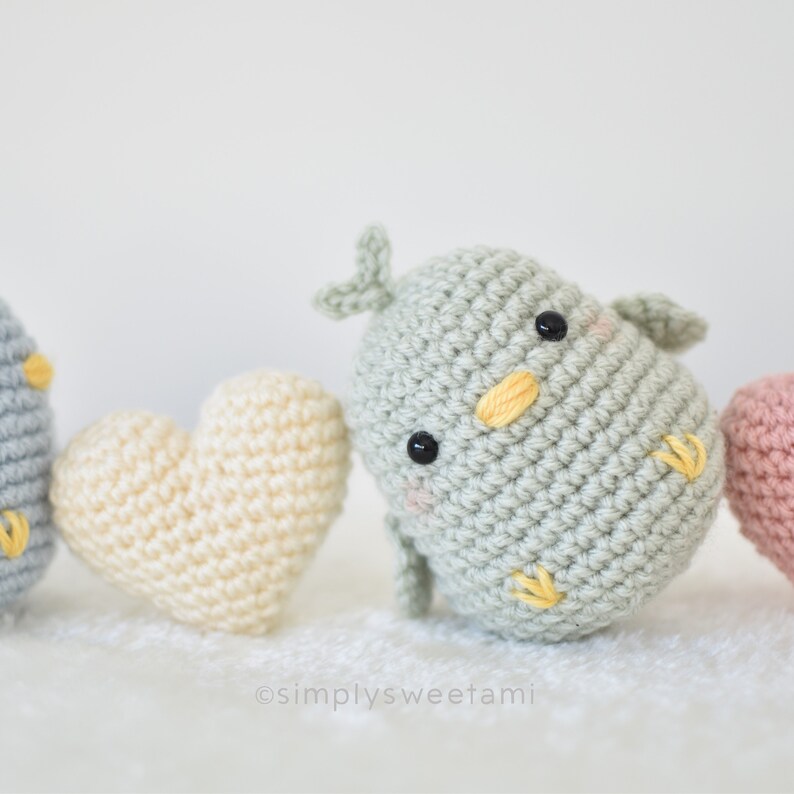 Może przedstawiać: Zestaw dzianych amigurumi ptak&oacute;w z ż&oacute;łtymi dziobami i czarnymi oczami, każdy siedzący obok dzianego serca. Serca są w kolorze kremowym, r&oacute;żowym i jasnoniebieskim.