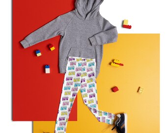 May include: A grey hooded sweatshirt with a white pair of leggings featuring a colourful retro boombox pattern. The leggings are paired with a pair of black trainers. The background is a red and yellow surface with small colourful toy blocks scattered around.