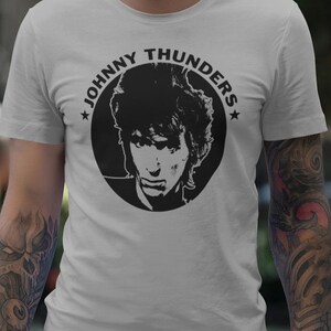 Puede incluir: Camiseta gris claro con un gráfico circular en blanco y negro de una cara y el texto "JOHNNY THUNDERS". La camiseta tiene mangas cortas.