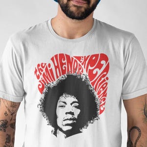 Op de afbeelding: Wit T-shirt met een zwart-wit portret van Jimi Hendrix en rode tekst erboven. De tekst luidt "Jimi Hendrix Experience" in een gestileerd lettertype. Het T-shirt heeft een ronde hals.