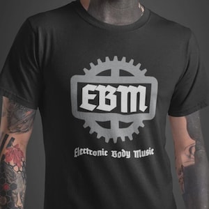 May include: Black t-shirt with a silver gear-shaped design and the letters "EBM" inside a rectangle. Below the design, the text reads "Electronic Body Music". The person in the image has tattoos and facial markings.