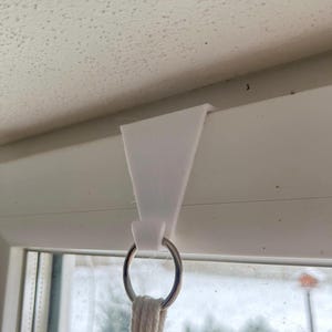 Könnte beinhalten: Weißer Plastikhaken, der an einem Fensterrahmen befestigt ist, mit einem silbernen Ring und einem weißen Seil, das daran hängt.