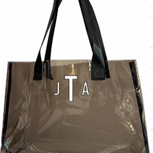 Może przedstawiać: Przezroczysta torba typu tote z czarnymi uchwytami i wykończeniami. Torba posiada biały monogram "JTA" na środku oraz napis "Monogram" w prawym g&oacute;rnym rogu. Torba wykonana jest z przezroczystego materiału.