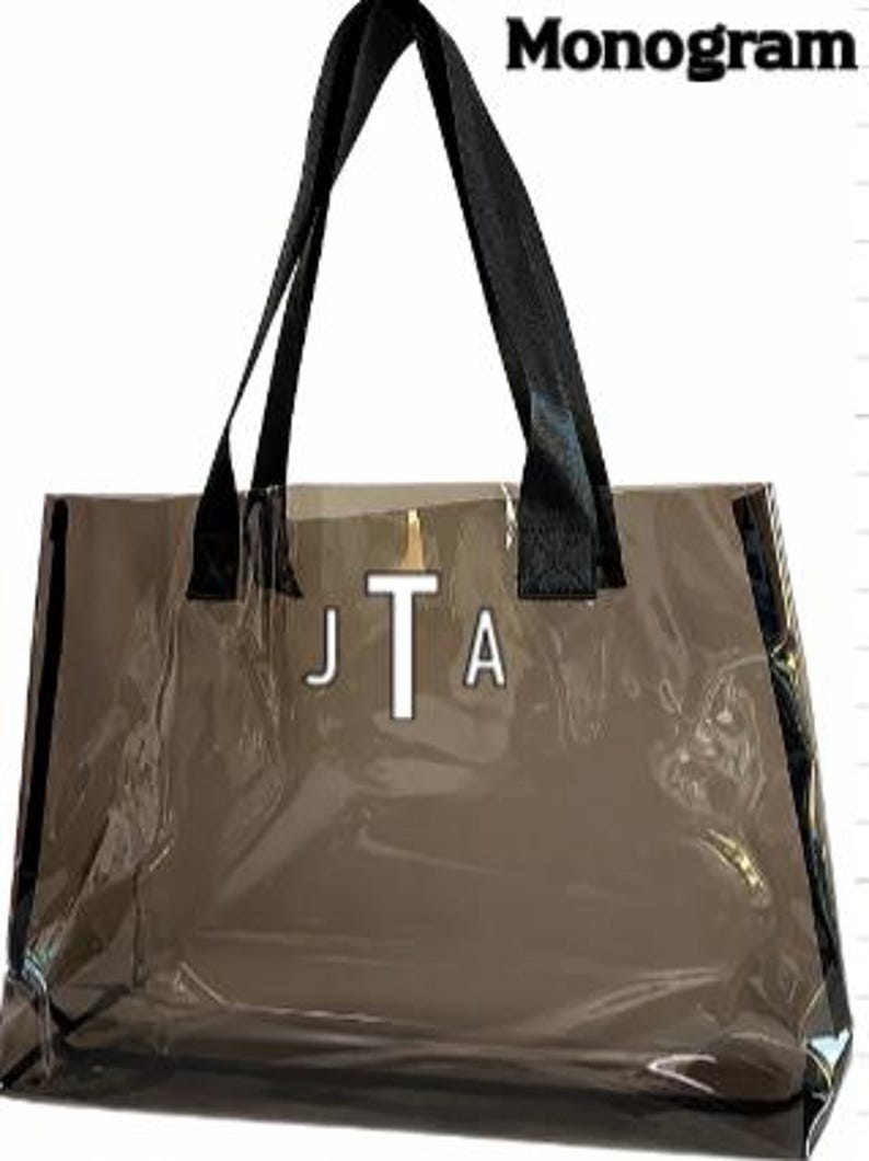 Może przedstawiać: Przezroczysta torba typu tote z czarnymi uchwytami i wykończeniami. Torba posiada biały monogram "JTA" na środku oraz napis "Monogram" w prawym g&oacute;rnym rogu. Torba wykonana jest z przezroczystego materiału.