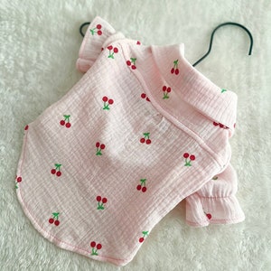 May include: A pink cotton shirt with a cherry print for a small dog. The shirt has a collar and short sleeves with ruffles.