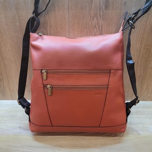May include: A burnt orange leather crossbody bag with two front zippered pockets and black adjustable straps. The bag has a rectangular shape and a top zipper closure. The bag is designed to be worn as a backpack or crossbody.
