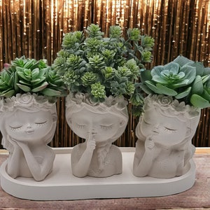 Può includere: Tre vasi in ceramica bianca a forma di volti di ragazze con gli occhi chiusi, ognuno con una pianta grassa. I vasi sono disposti su un vassoio ovale bianco.